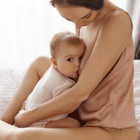 Young tender mother in sleepwear breastfeeding hugging her newborn baby sitting in bed at morning. Copy space.